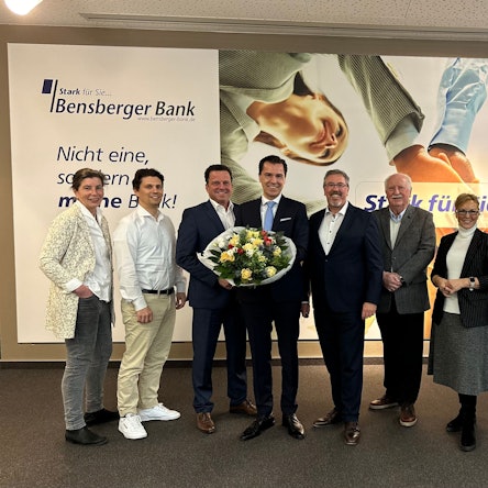 Vorstand und Aufsichtsrat der Bensberger Bank stehen vor einer Reklame der Genossenschaftsbank.