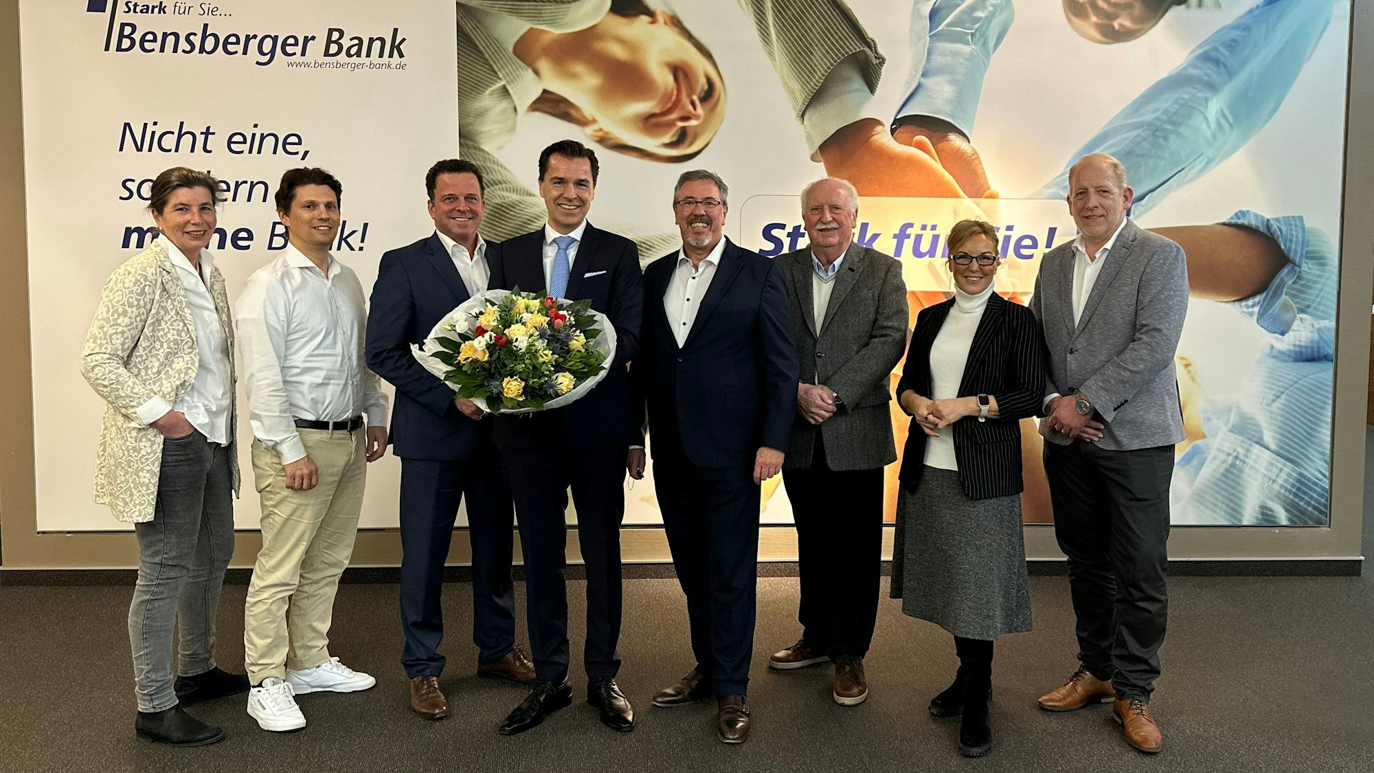 Vorstand und Aufsichtsrat der Bensberger Bank stehen vor einer Reklame der Genossenschaftsbank.