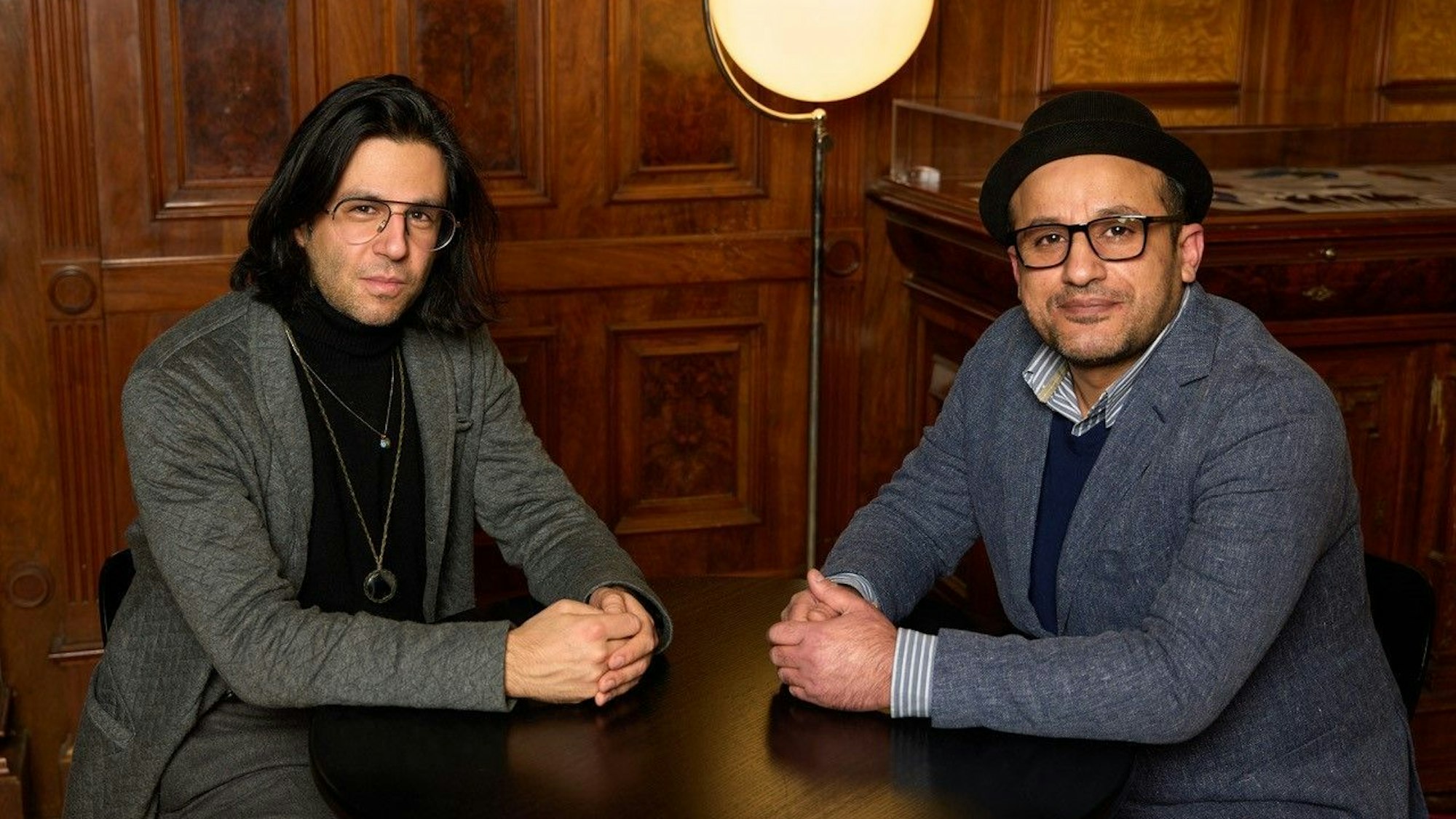 Deniz Utlu und Reber Yousef sitzen beide an einem kleinen Tisch und schauen in die Kamera.Sie haben die Hände ineinander verschränkt und auf dem Tisch angelehnt. Im Hintergrund eine holzvertäfelte Wand und eine Lampe.