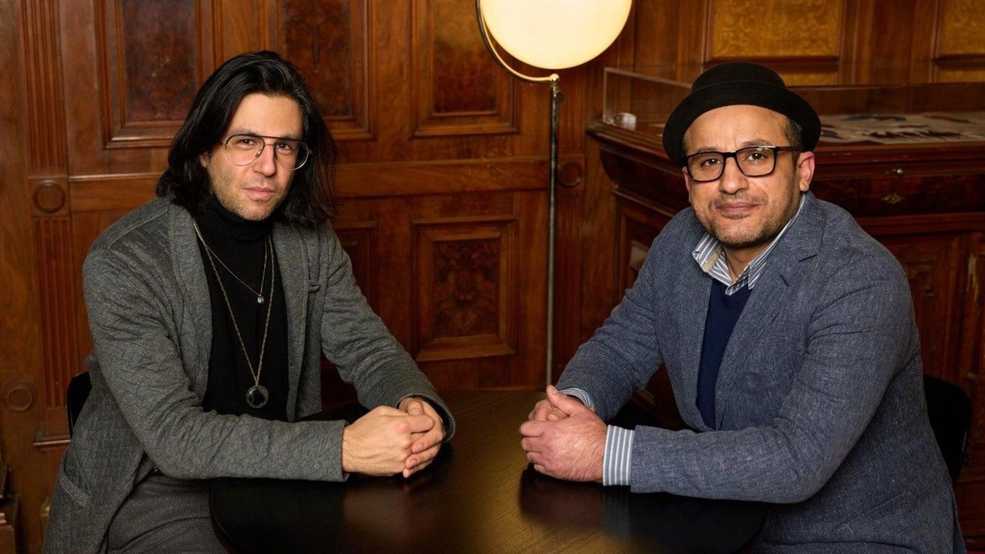 Deniz Utlu und Reber Yousef sitzen beide an einem kleinen Tisch und schauen in die Kamera.Sie haben die Hände ineinander verschränkt und auf dem Tisch angelehnt. Im Hintergrund eine holzvertäfelte Wand und eine Lampe.