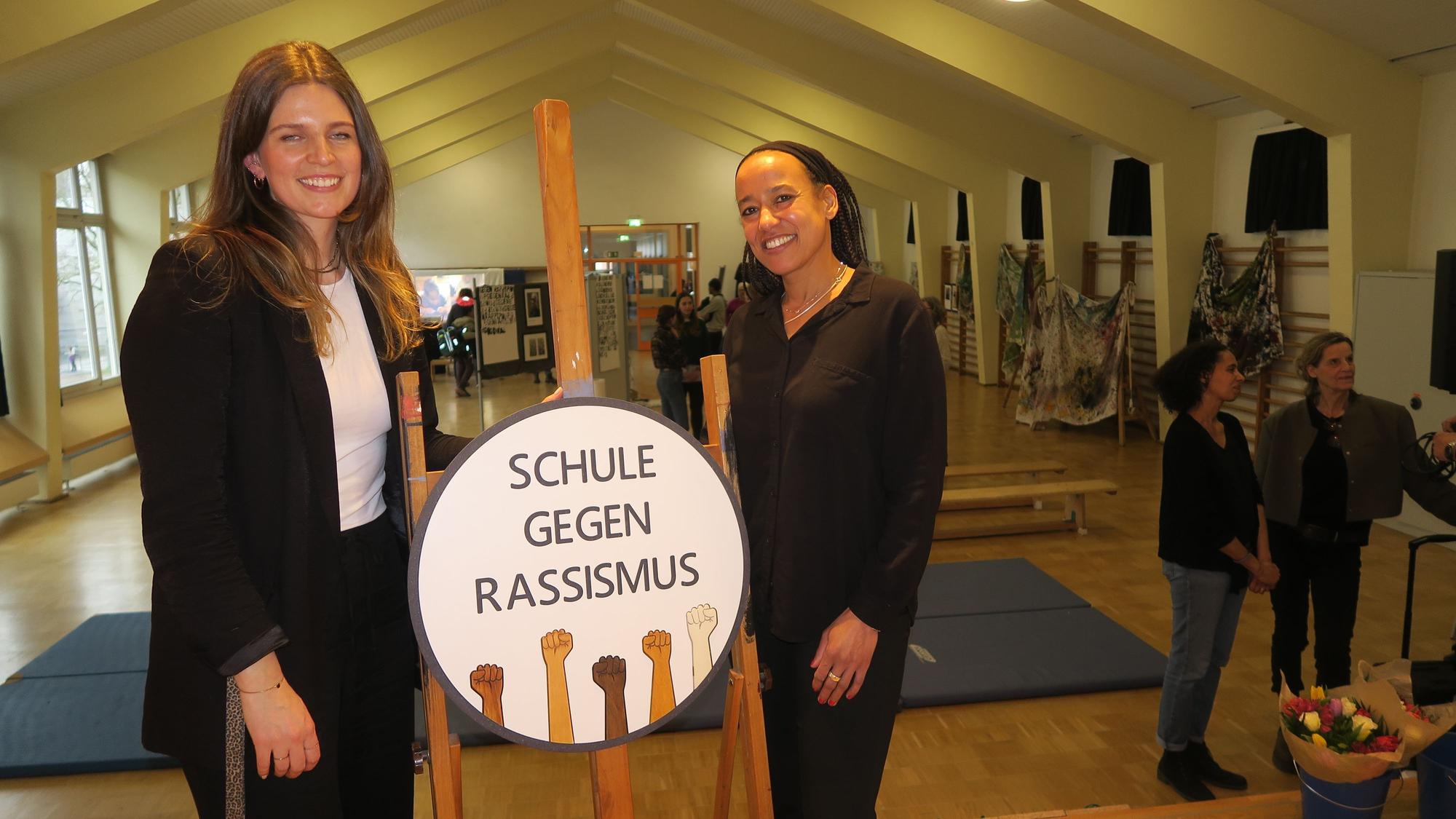 In einer Turnhalle stehen zwei junge Frauen bei einem Aufsteller, auf dem Schule gegen Rassismus steht