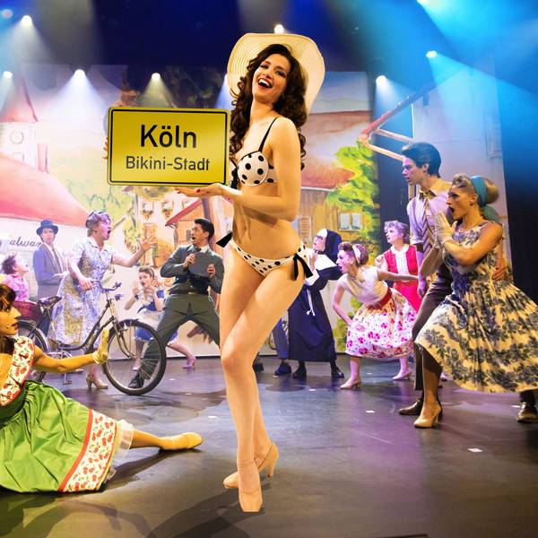 Eine Frau im Bikini hält ein Schild mit der Aufschrift „Köln – Bikini-Stadt“. Das Musical „Bikini Skandal“ kommt nach Köln.