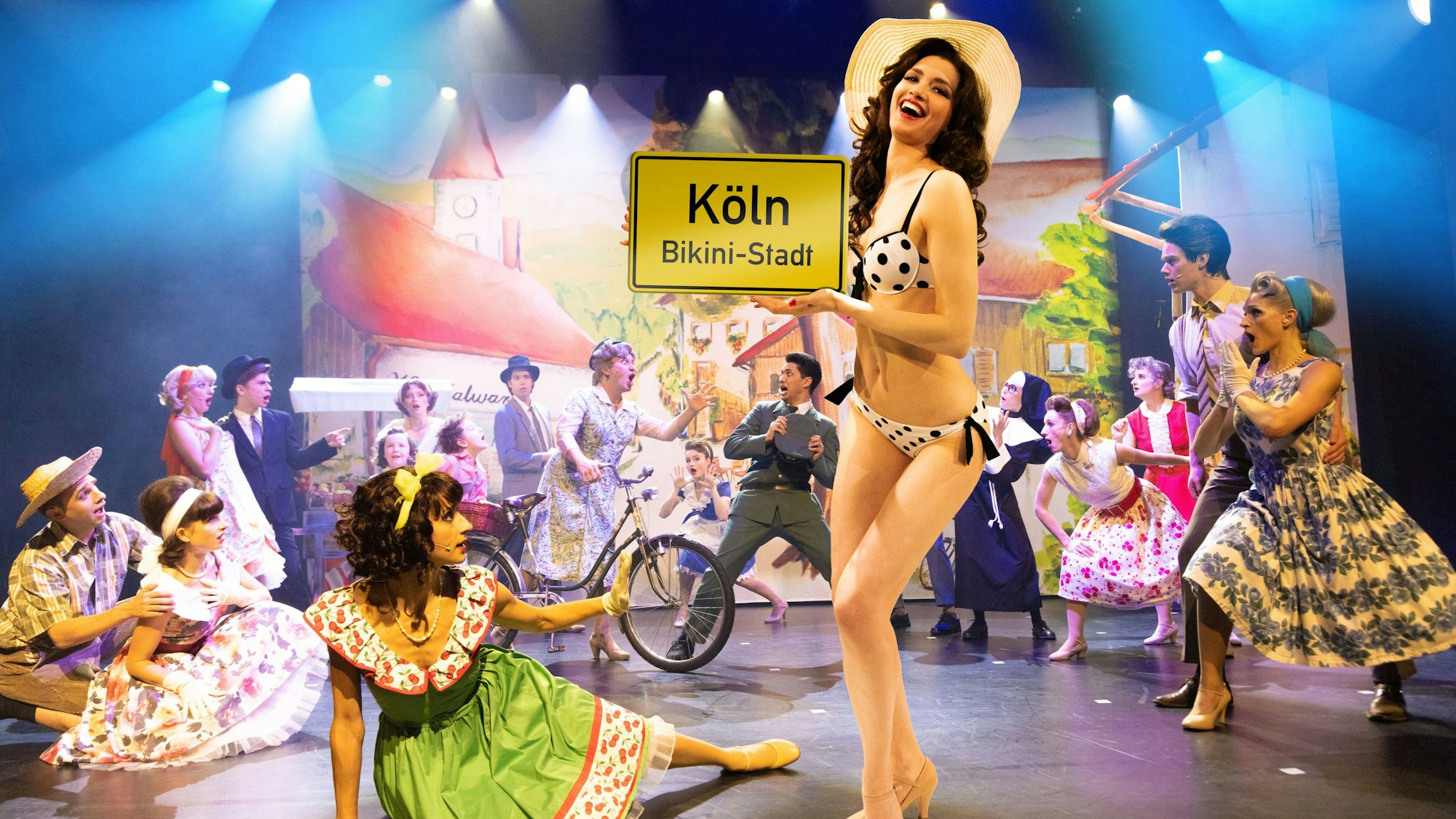 Eine Frau im Bikini hält ein Schild mit der Aufschrift „Köln – Bikini-Stadt“. Das Musical „Bikini Skandal“ kommt nach Köln.