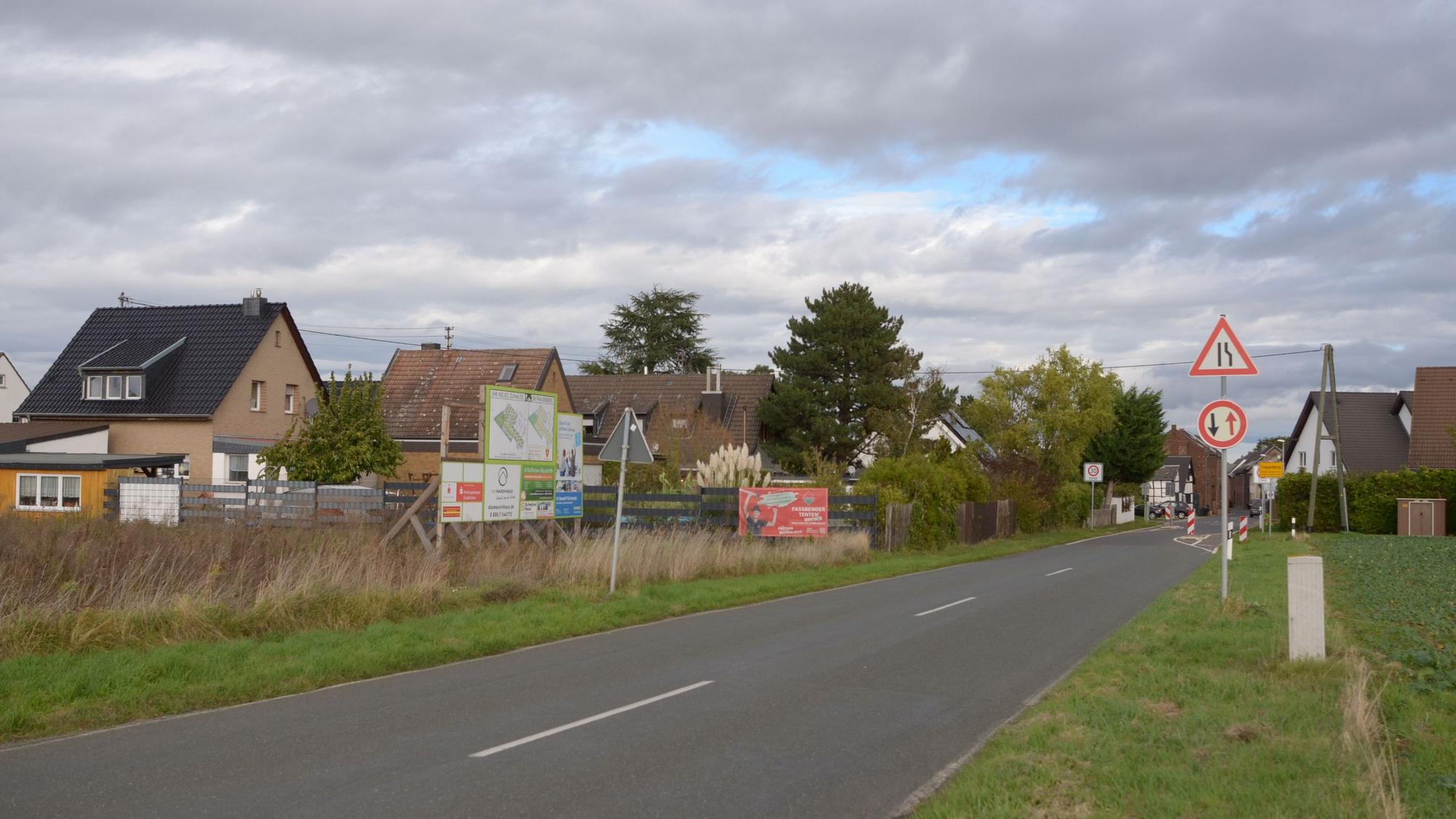 Das Bild zeigt die Nideggener Straße, die nach Frauenberger hineinführt. Am Straßenrand steht eine Werbetafel, die auf das geplante Wohngebiet aufmerksam macht.