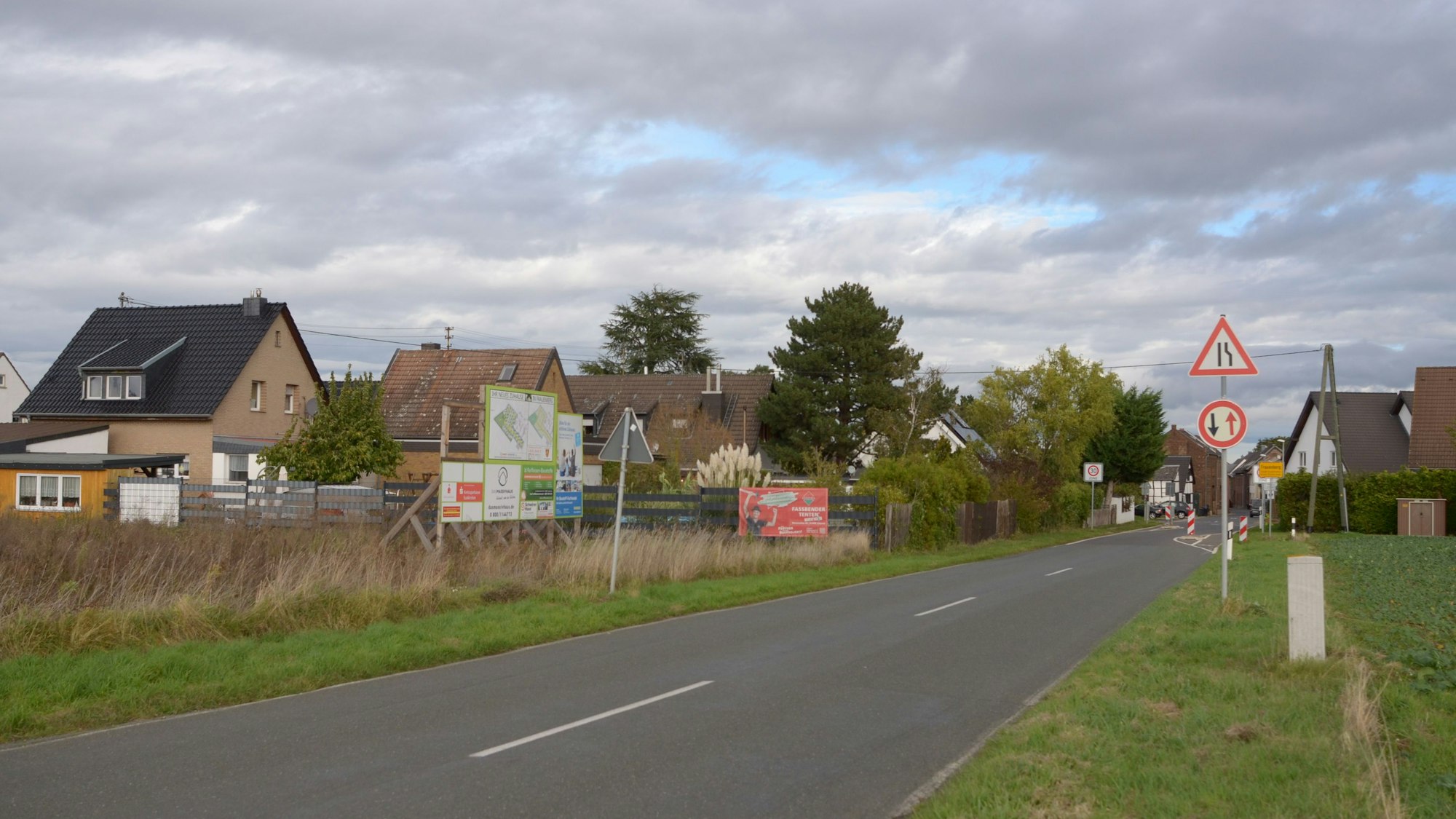 Das Bild zeigt die Nideggener Straße, die nach Frauenberger hineinführt. Am Straßenrand steht eine Werbetafel, die auf das geplante Wohngebiet aufmerksam macht.