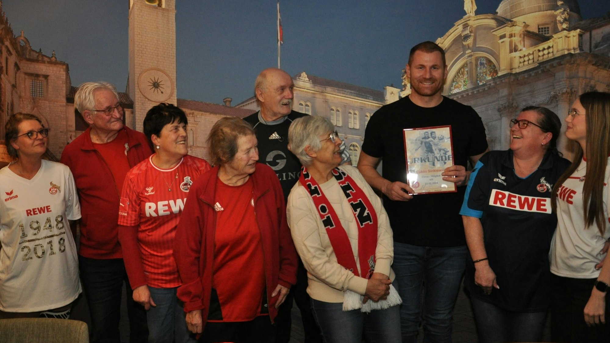 Der ehemalige Kapitän des 1. FC Köln, Lukas Sinkiewicz, hält eine Urkunde in der Hand. Einige Frauen und Männer in FC-Trikots schauen ihn an.