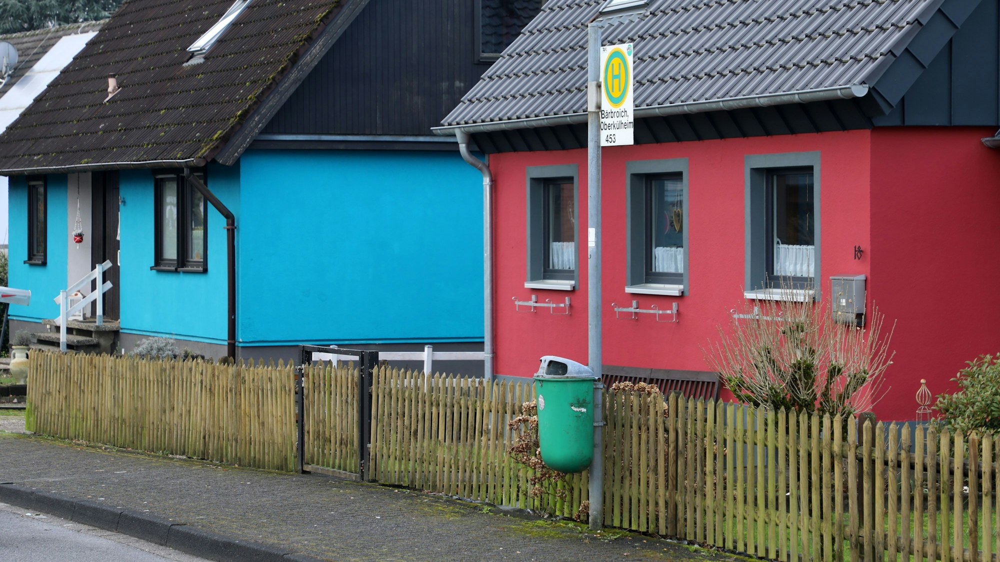 Die einsamste Bushaltestelle Bergisch Gladbachs steht in Oberkühlheim. Ein Mülleimer und ein Bushaltestellenschild stehen vor einem roten Haus, weiter hinten links im Bild ist ein türkises Haus.