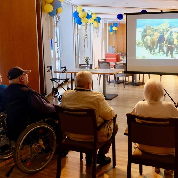 Einige ältere Menschen sitzen im Halbkreis in einem großen Saal. Sie betrachten ein Bild, das auf eine Leinwand projiziert wird.