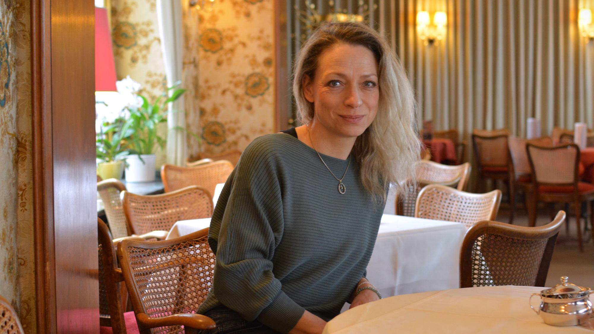 Carolin Soyka sitzt in einem Café, sie hat mittellange blonde Haare und trägt einen blaugrauen Pullover.
