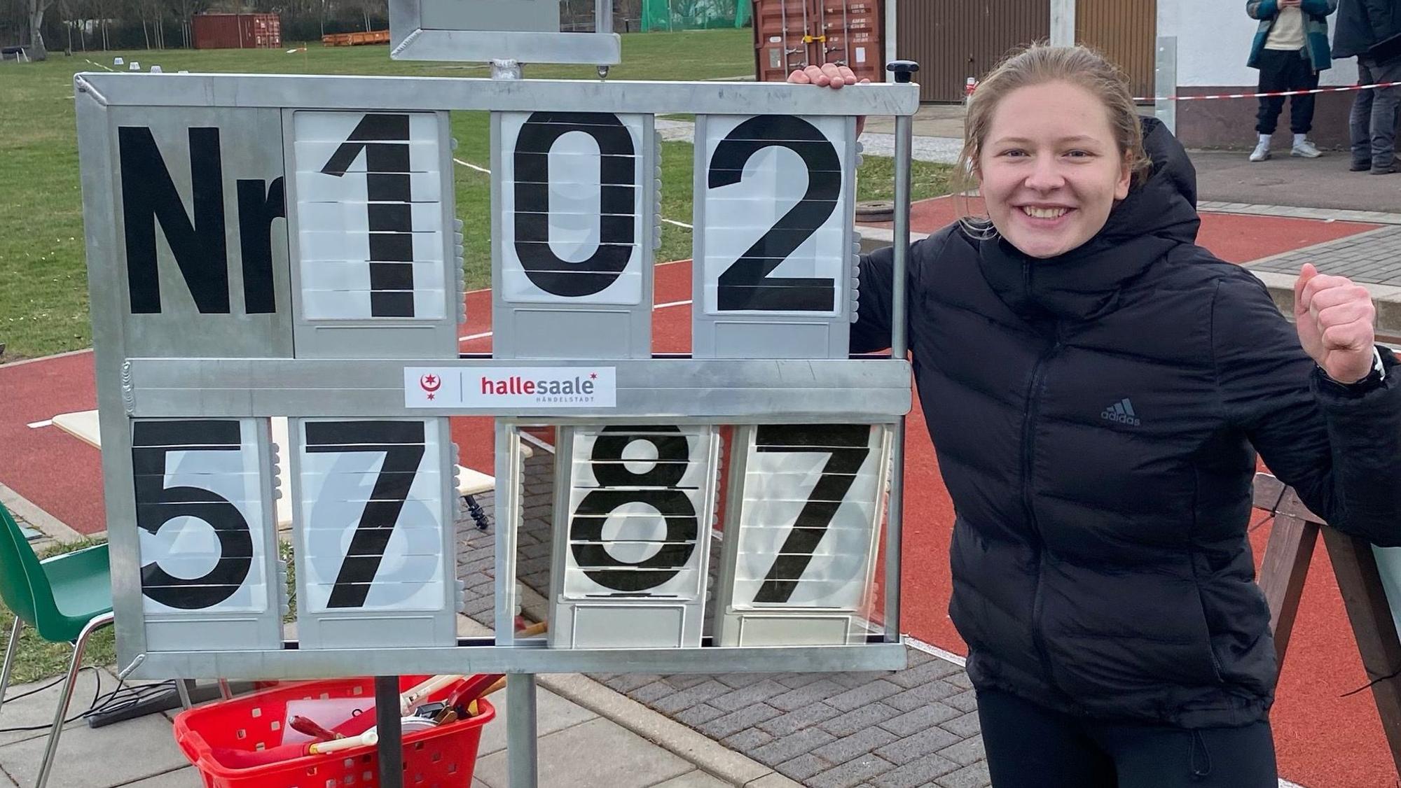 Victoria Krause steht neben der Anzeigentafel, auf der die Siegesweite von 57,87 Meter abgebildet ist und ballt ihre Faust.