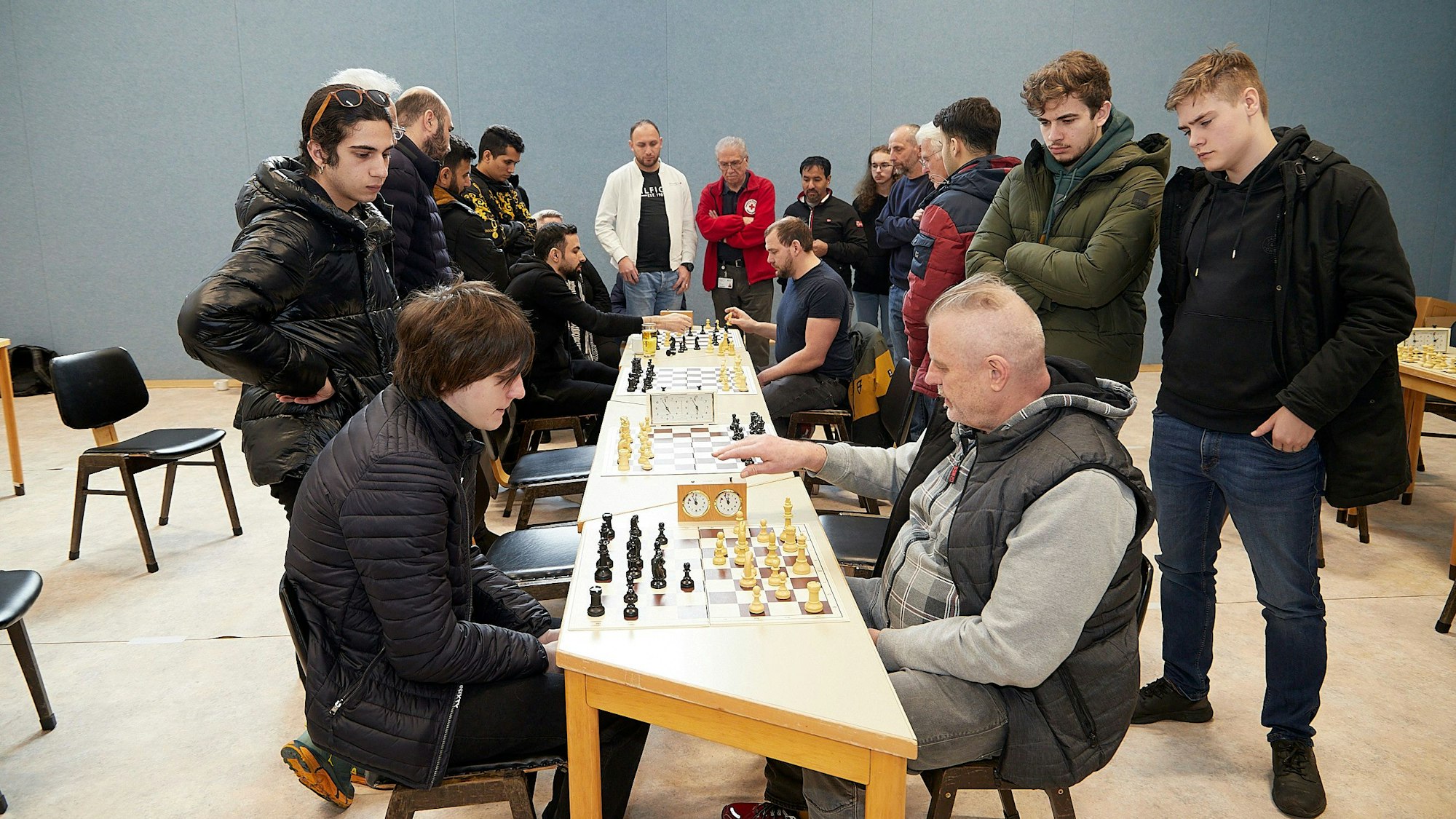 An zwei Schachtischen sitzen sich die Spieler gegenüber. Eine Gruppe Menschen steht um sie herum und schaut ihnen zu.