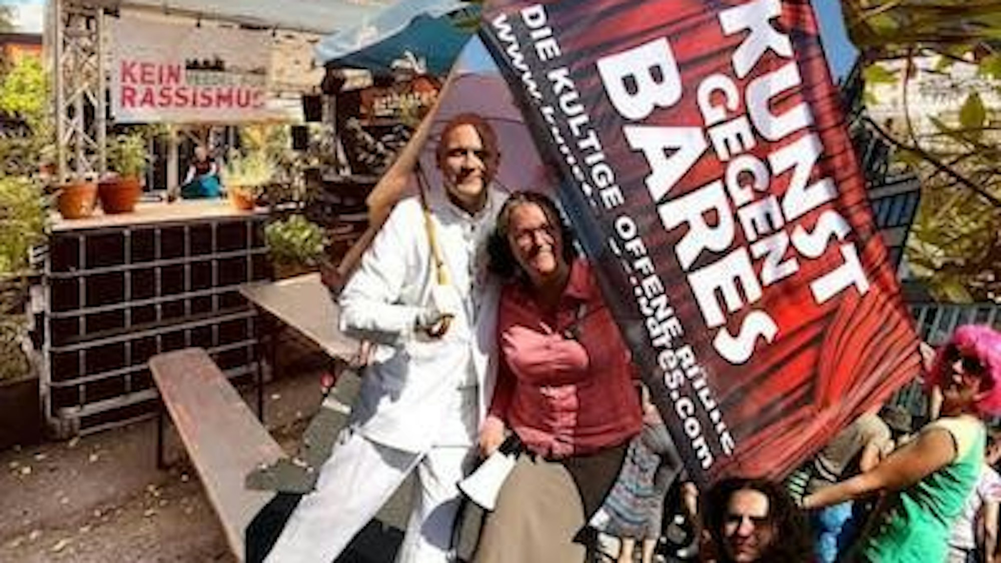 Auf dem Foto sieht man Gerd Buurmann sowie andere Menschen, die eine Flagge in der Hand halten, auf der „Kunst gegen Bares“ steht.