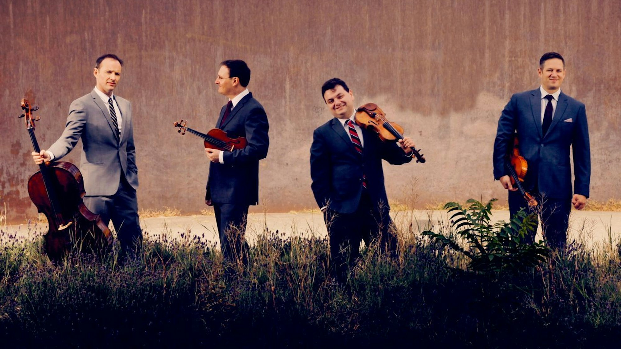 Das Bild zeigt die vier Musiker vor einer Wand im hohen Gras stehen. Alle halten ihre Streichinstrumente in der Hand. Der Cellist trägt einen grauen Anzug, die anderen blaue.