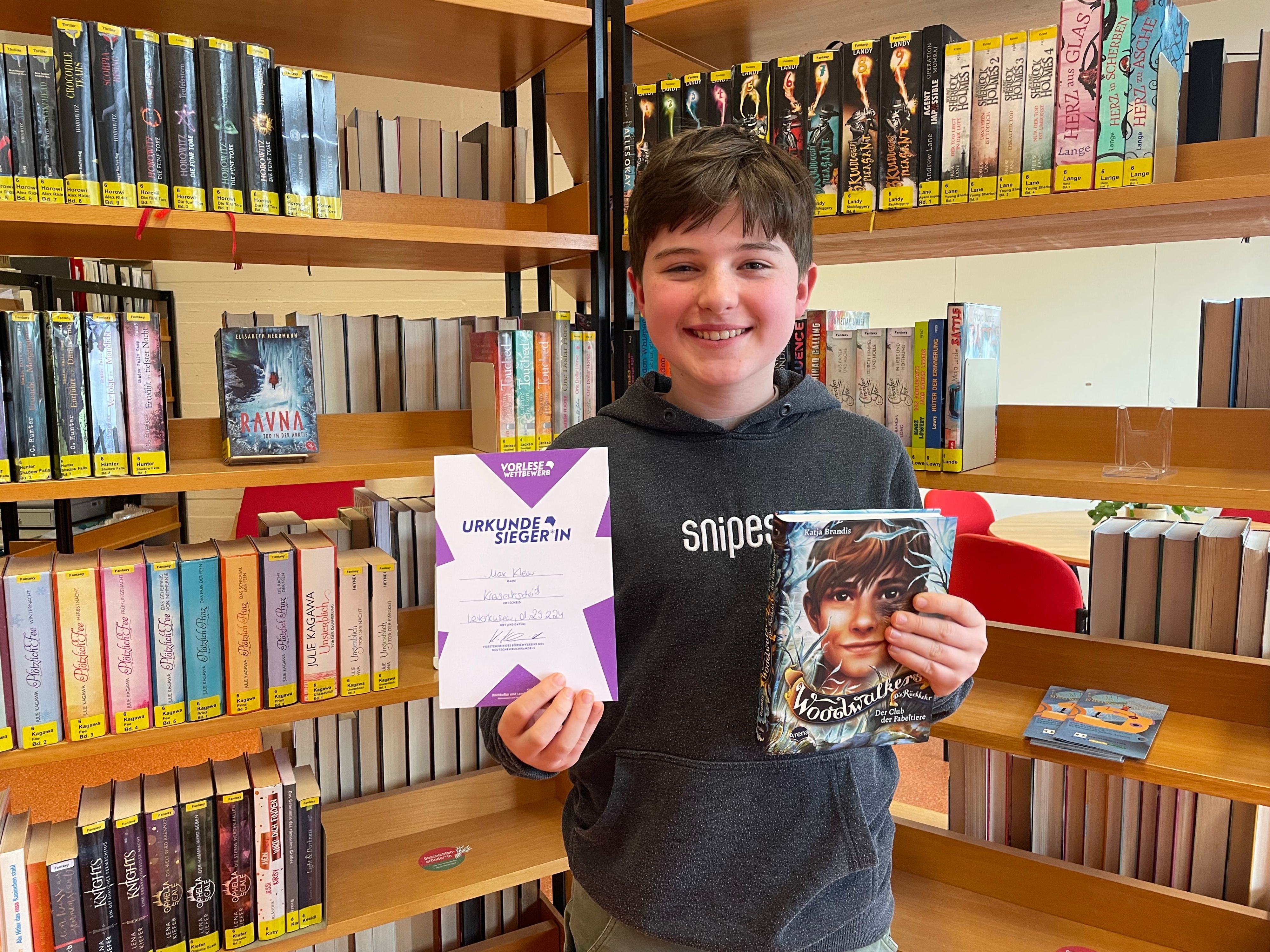 Max Klein steht vor einem Buchregal, hält seine Siegerurkunde in der einen und das Buch „Woodwalkers“ in der anderen Hand und lächelt in die Kamera.