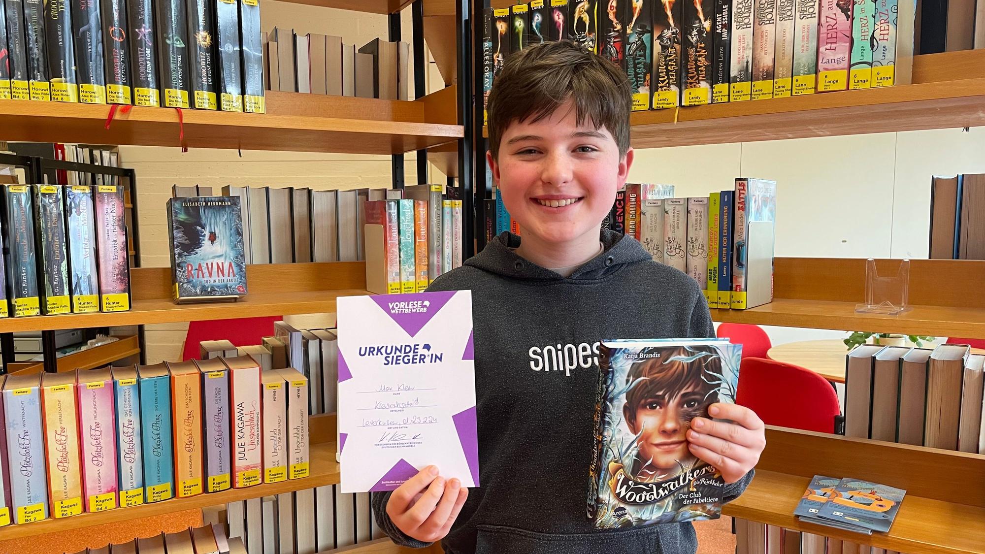 Max Klein steht vor einem Buchregal, hält seine Siegerurkunde in der einen und das Buch „Woodwalkers“ in der anderen Hand und lächelt in die Kamera.