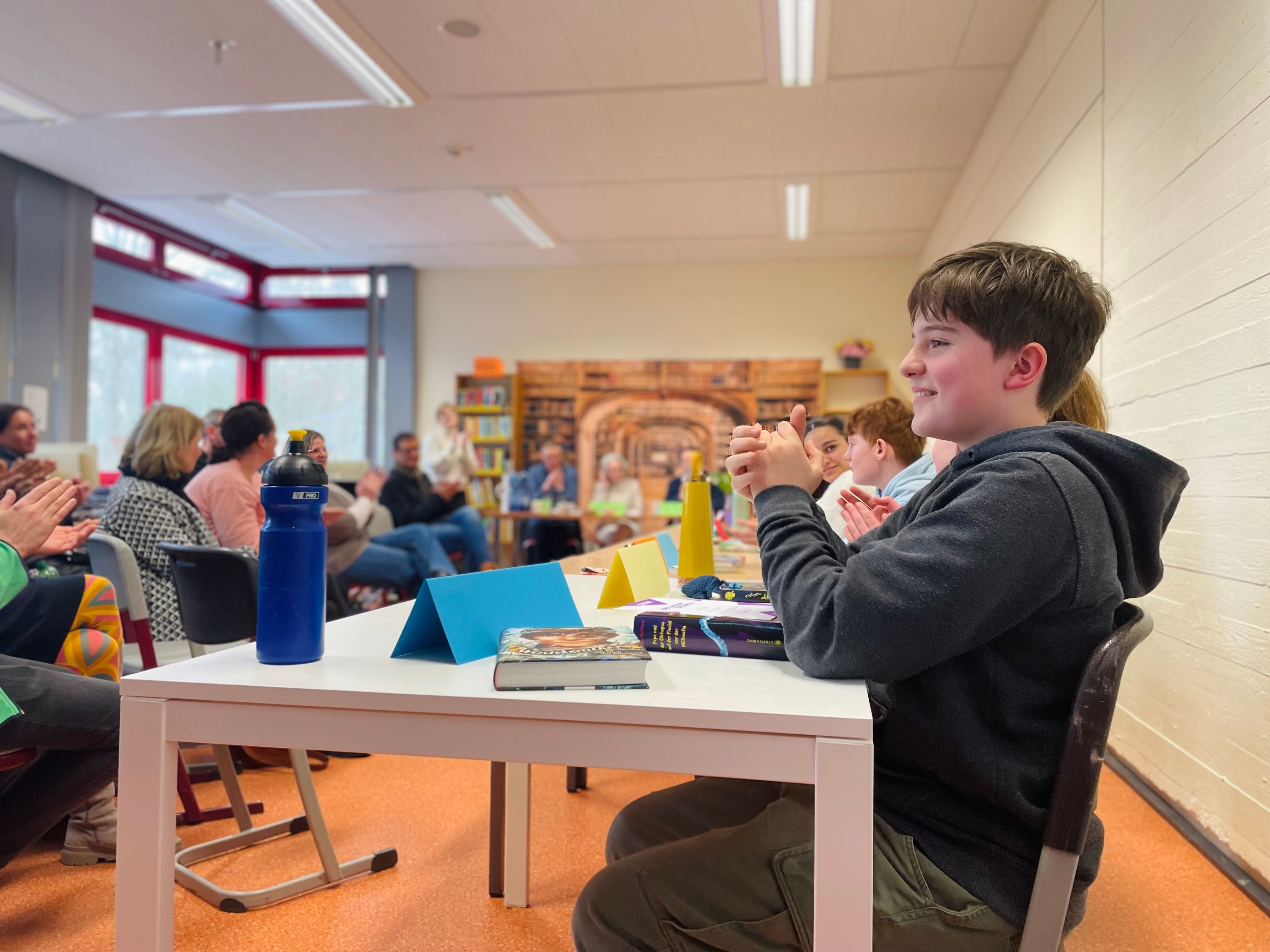 Max Klein sitzt am Tisch und klatscht, zwei Bücher liegen vor ihm. Im Hintergrund sind weitere Schülerinnen und Schüler des Vorlesewettbewerbs, sowie das Publikum zu sehen.