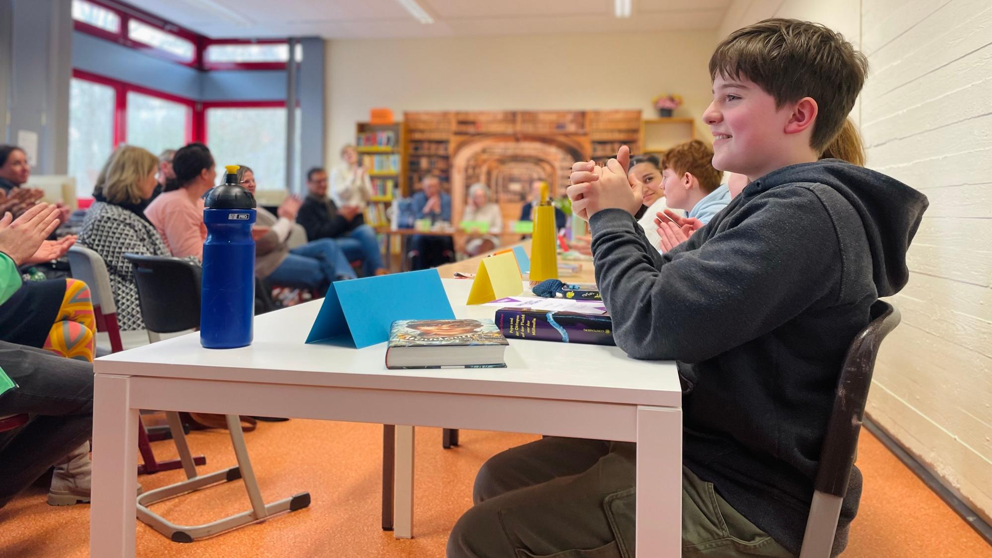 Max Klein sitzt am Tisch und klatscht, zwei Bücher liegen vor ihm. Im Hintergrund sind weitere Schülerinnen und Schüler des Vorlesewettbewerbs, sowie das Publikum zu sehen.