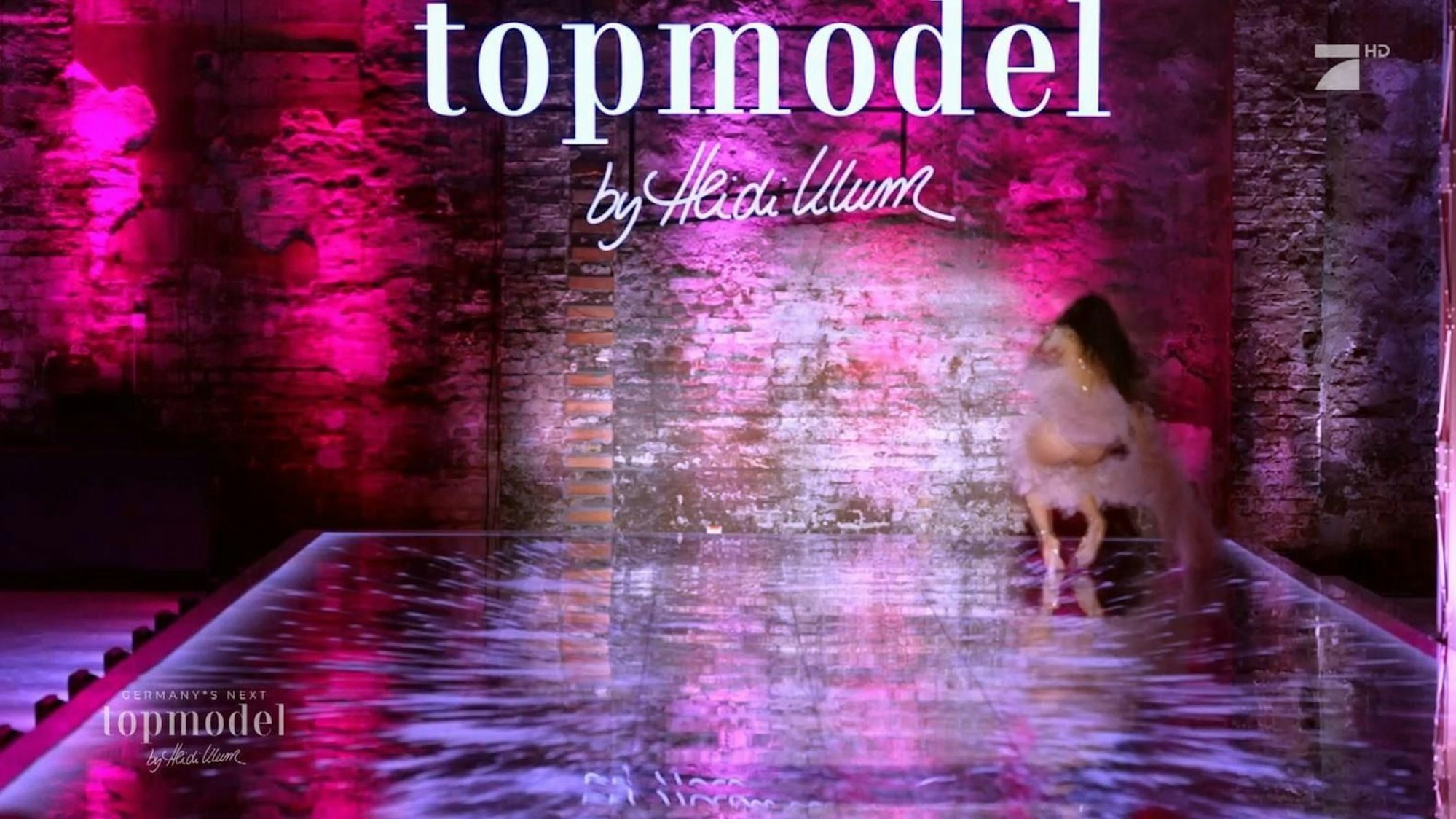 Hinten rechts stürzt Topmodel-Anwärterin in der dritten Folge von GNTM 2024 auf einem lila beleuchteten Catwalk.