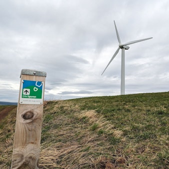 Ein Markierungspfosten eines Wanderwegs steht neben einer Wiese, auf dem sich ein Windrad und die Reste eines Flakbunkers aus dem Zweiten Weltkrieg befinden.