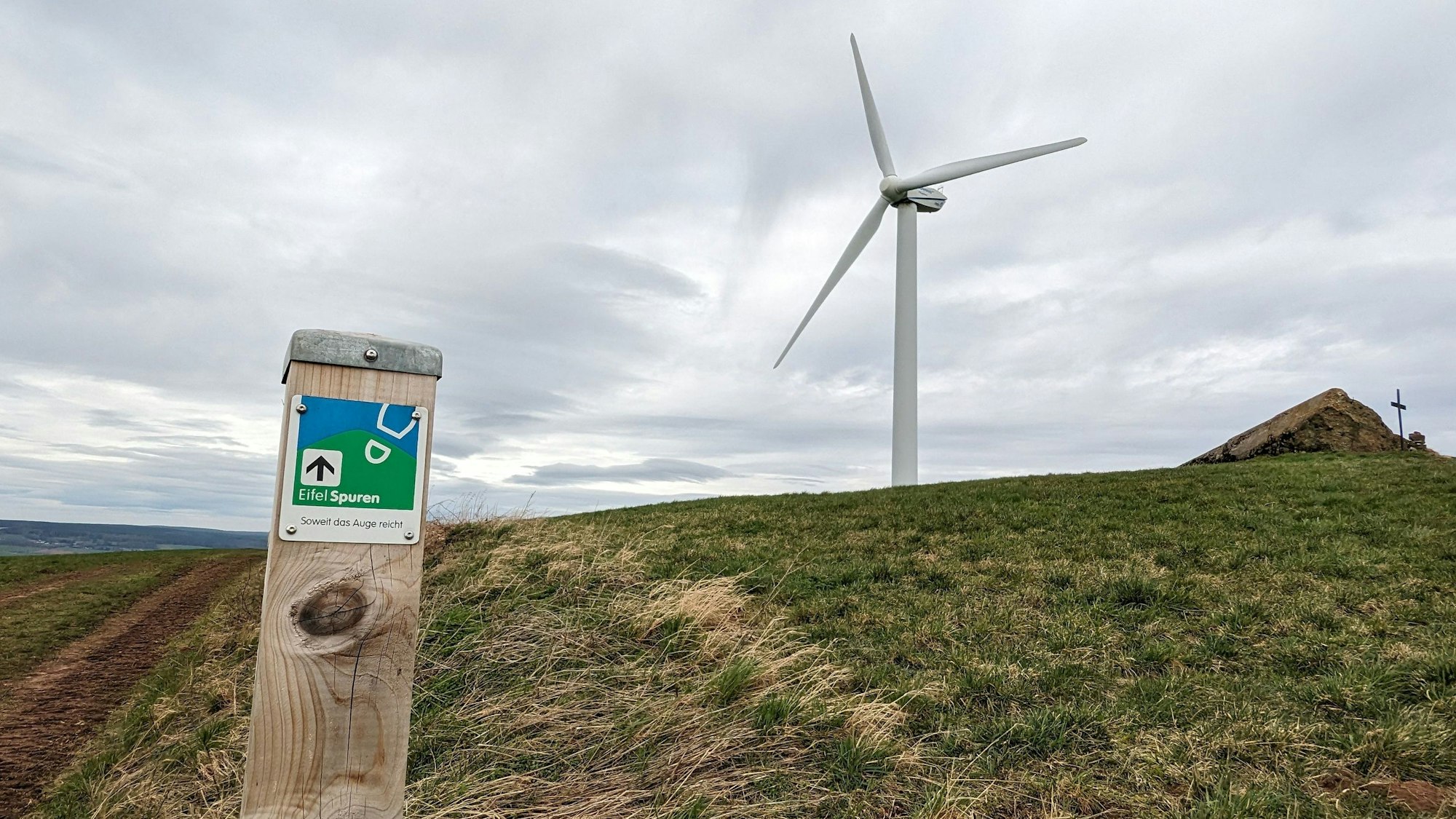 Ein Markierungspfosten eines Wanderwegs steht neben einer Wiese, auf dem sich ein Windrad und die Reste eines Flakbunkers aus dem Zweiten Weltkrieg befinden.