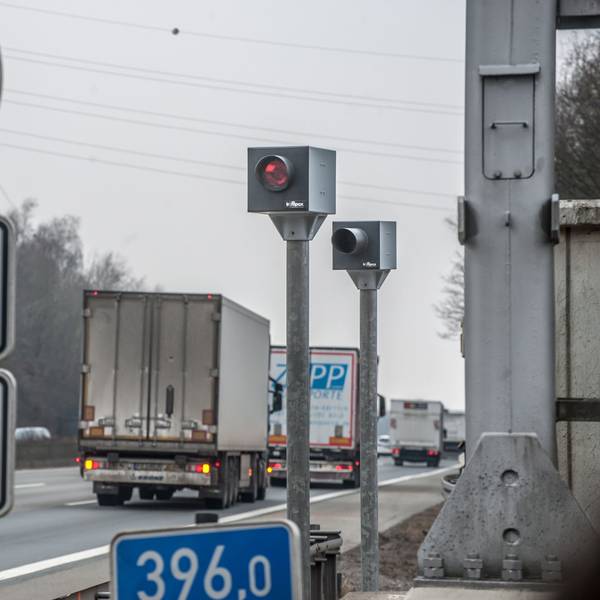 Ein Blitzer steht an einer Autobahn. An ihm fahren Lkws und Pkws vorbei. Vorne links steht ein Straßenschild.
