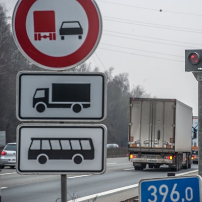 Ein Blitzer steht an einer Autobahn. An ihm fahren Lkws und Pkws vorbei. Vorne links steht ein Straßenschild.