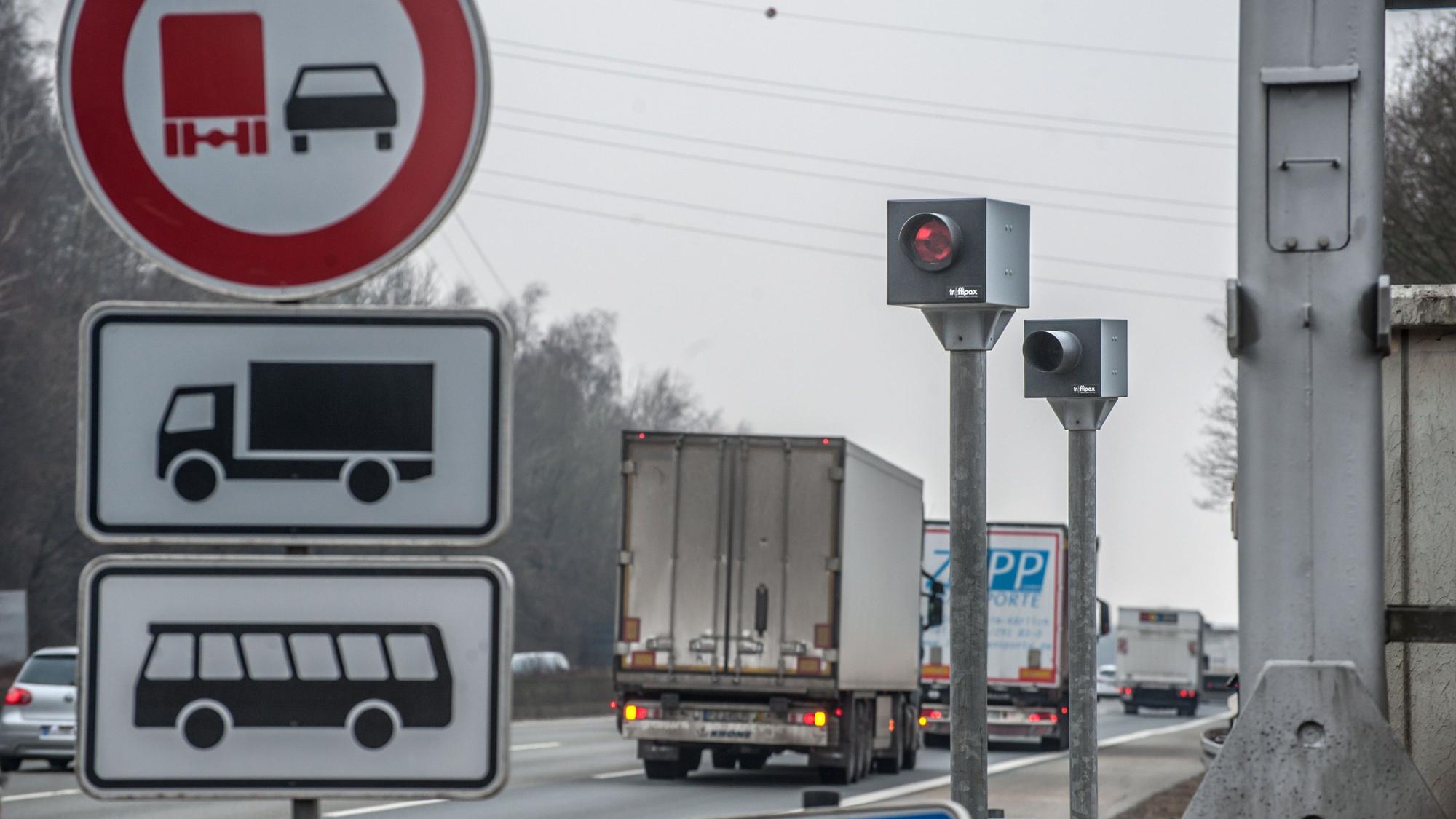 Ein Blitzer steht an einer Autobahn. An ihm fahren Lkws und Pkws vorbei. Vorne links steht ein Straßenschild.