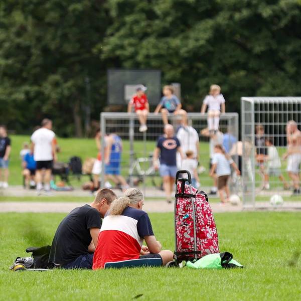 Viele Kinder und Jugendliche sitzen auf einer Wiese, im Hintergrund spielen einige Fußball auf einem Bolzplatz.