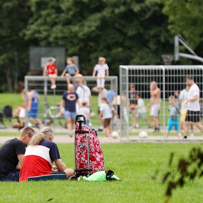 Viele Kinder und Jugendliche sitzen auf einer Wiese, im Hintergrund spielen einige Fußball auf einem Bolzplatz.