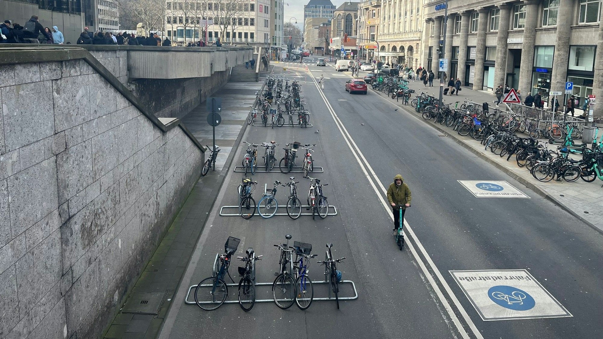 Das Bild zeigt eine Straße am Kölner Dom mit Fahrradständern und Menschen.