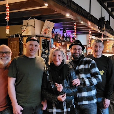 Eine Gruppe von gut gelaunten Männern und Frauen vor einer Theke im Schankraum einer Kneipe.