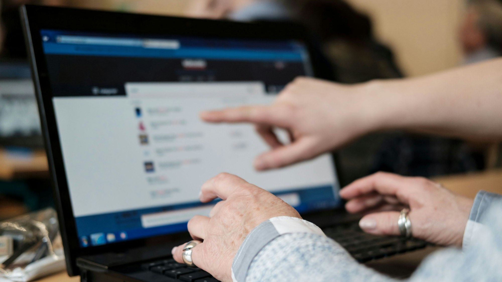 Eine Teilnehmerin des Computerkurses für Senioren übt zusammen mit einem Schüler an einem Laptop. Sie hält ihre Hände über die Tastatur, während ein Schüler mit einem Finger auf den Bildschirm zeigt.