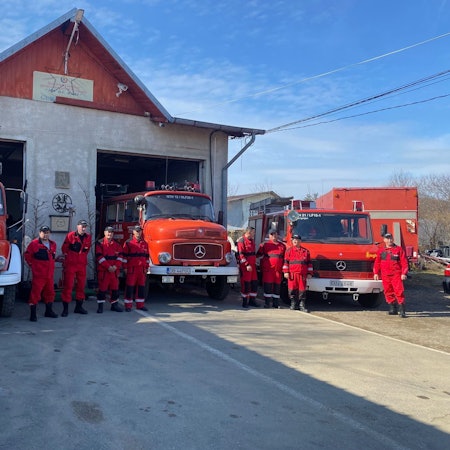 Rot gekleidete Feuerwehrleute stehen vor drei roten Feuerwehrwagen.