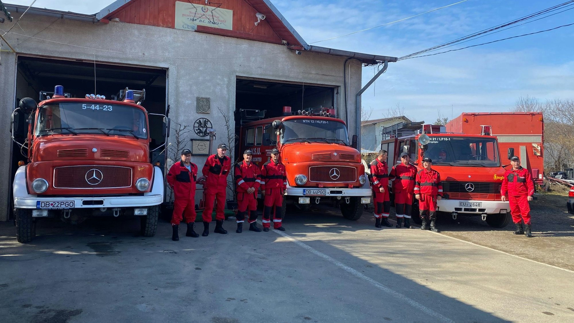 Rot gekleidete Feuerwehrleute stehen vor drei roten Feuerwehrwagen.