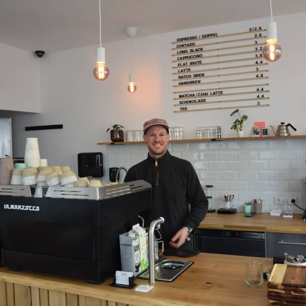 Ein junger Mann mit Kappe steht hinter eine Kaffeemaschine in seinem Café.