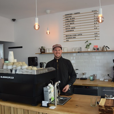 Ein junger Mann mit Kappe steht hinter eine Kaffeemaschine in seinem Café.