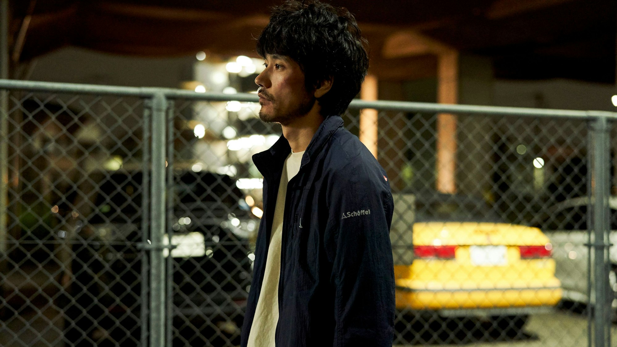 Auf dem Bild ist Ken'ichi Matsuyama in seiner Rolle als Uri zu sehen. Er wirkt niedergeschlagen. Im Hintergrund ist verschwommen ein Zaun zu sehen, dahinter parken Autos.