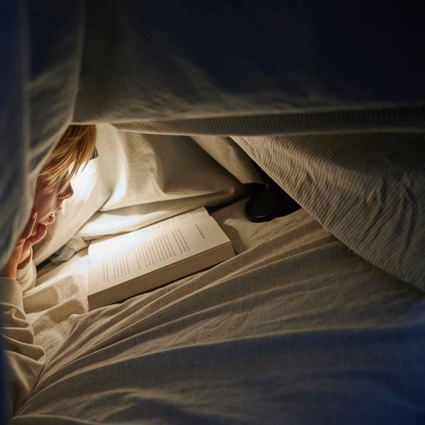 Ein Kind liegt auf einem Bett in einem dunklen Zimmer und liest unter einer Bettdecke mit einer Lampe ein Buch.