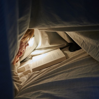 Ein Kind liegt auf einem Bett in einem dunklen Zimmer und liest unter einer Bettdecke mit einer Lampe ein Buch.