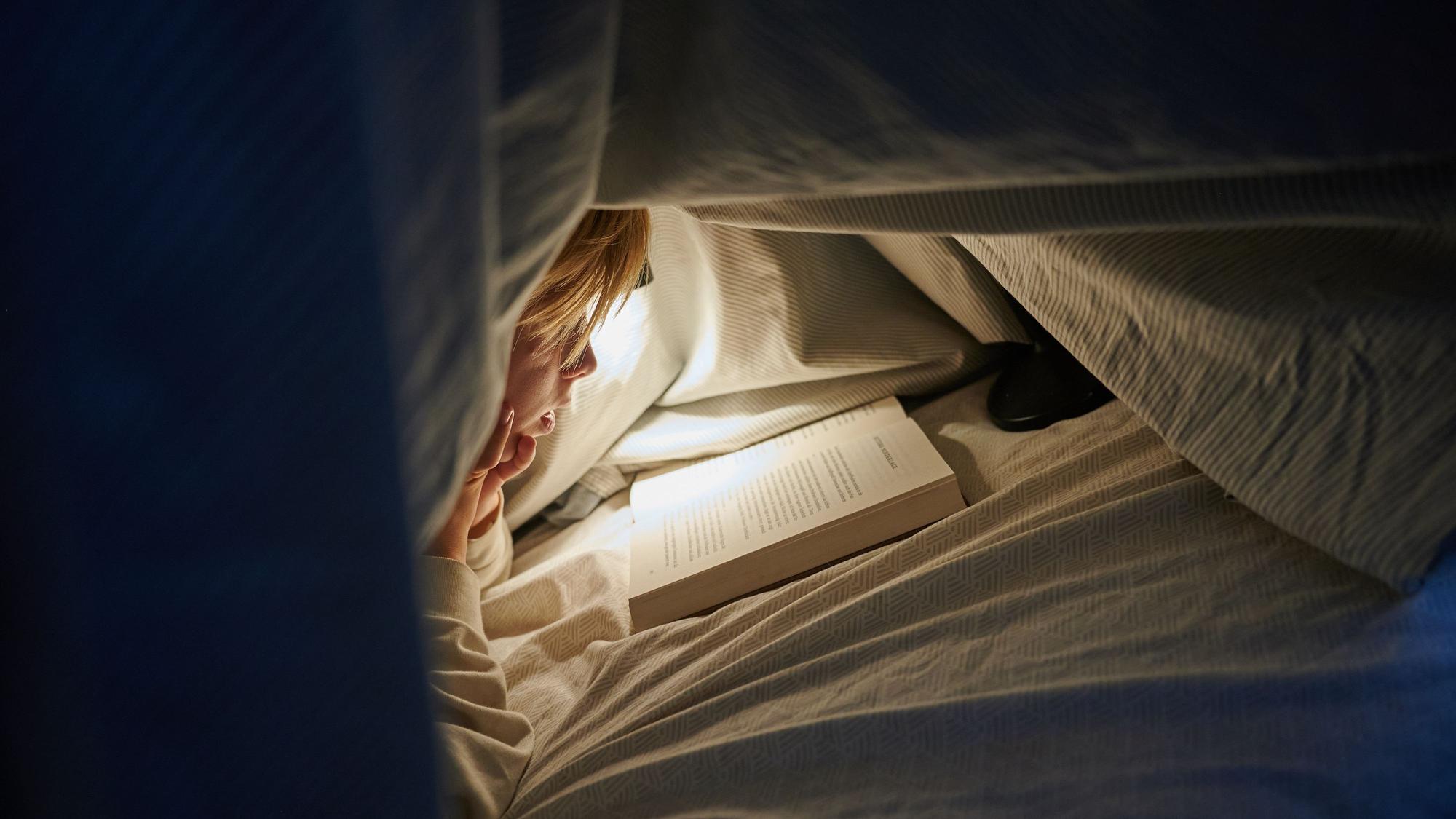 Ein Kind liegt auf einem Bett in einem dunklen Zimmer und liest unter einer Bettdecke mit einer Lampe ein Buch.
