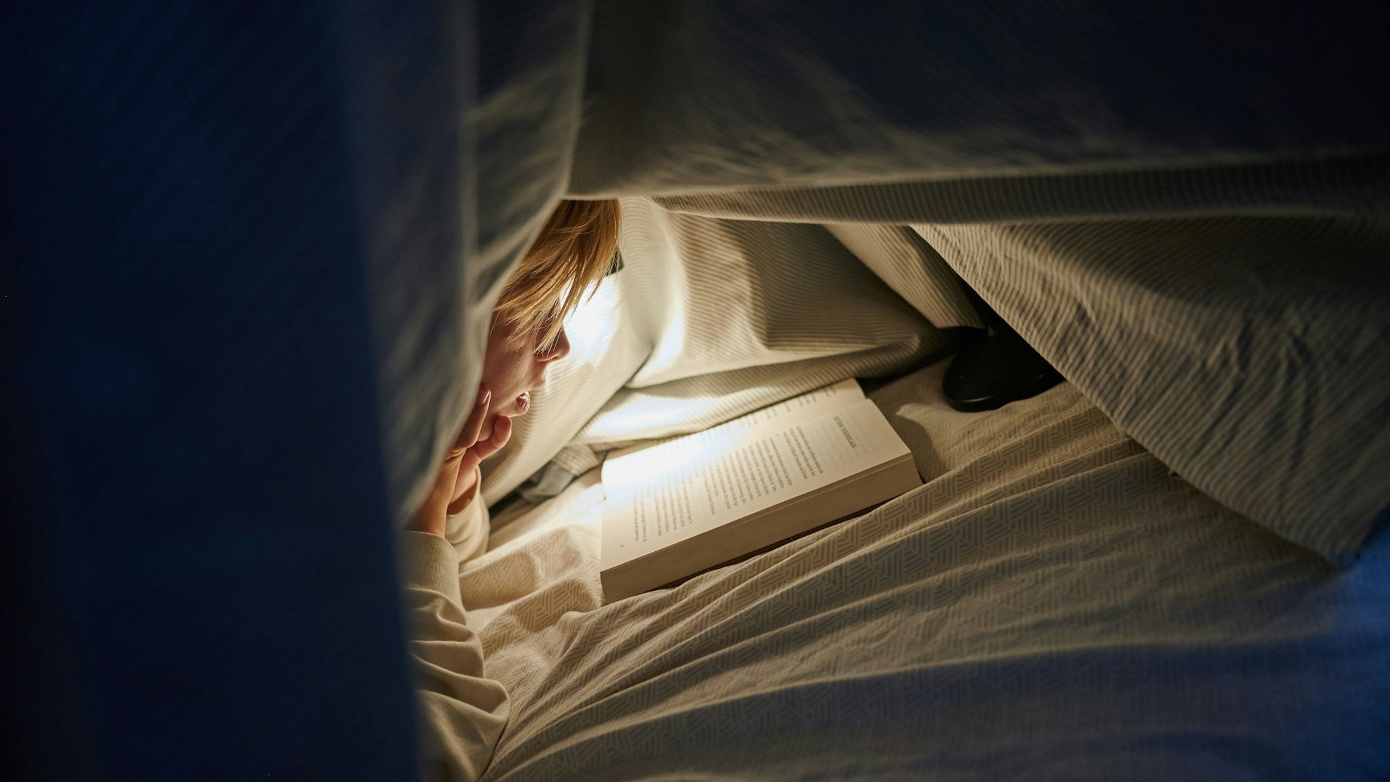 Ein Kind liegt auf einem Bett in einem dunklen Zimmer und liest unter einer Bettdecke mit einer Lampe ein Buch.