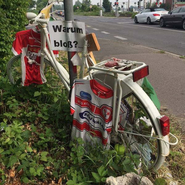 Ein weißes Fahrrad mit persönlichen Gegenständen wir Trikots steht in der Nähe der Unfallstelle am Straßenrand.