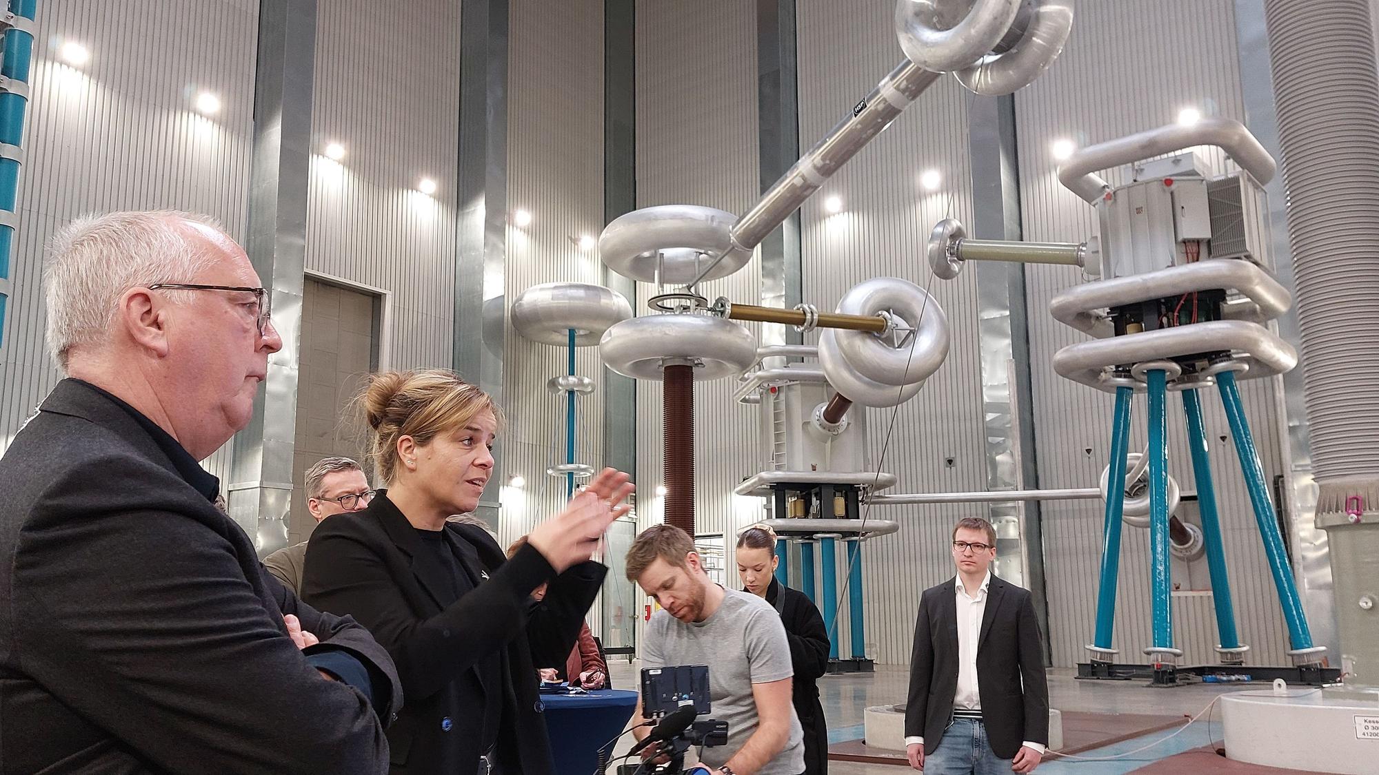 Eine blonde Frau in schwarzer Kleidung spricht in einer Industriehalle. Im Hintergrund stehen Geräte für Hochspannungsprüfungen.