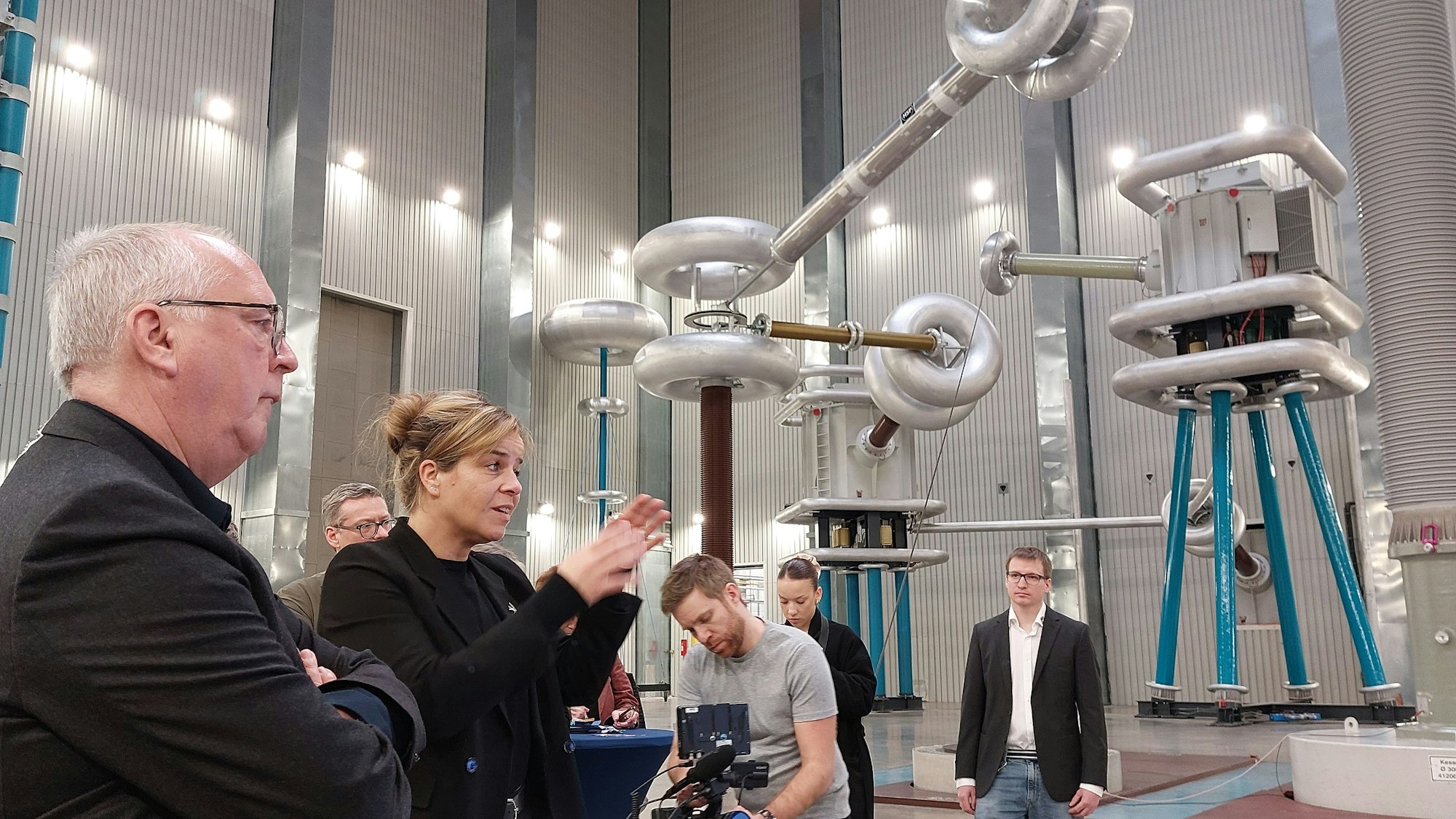 Eine blonde Frau in schwarzer Kleidung spricht in einer Industriehalle. Im Hintergrund stehen Geräte für Hochspannungsprüfungen.