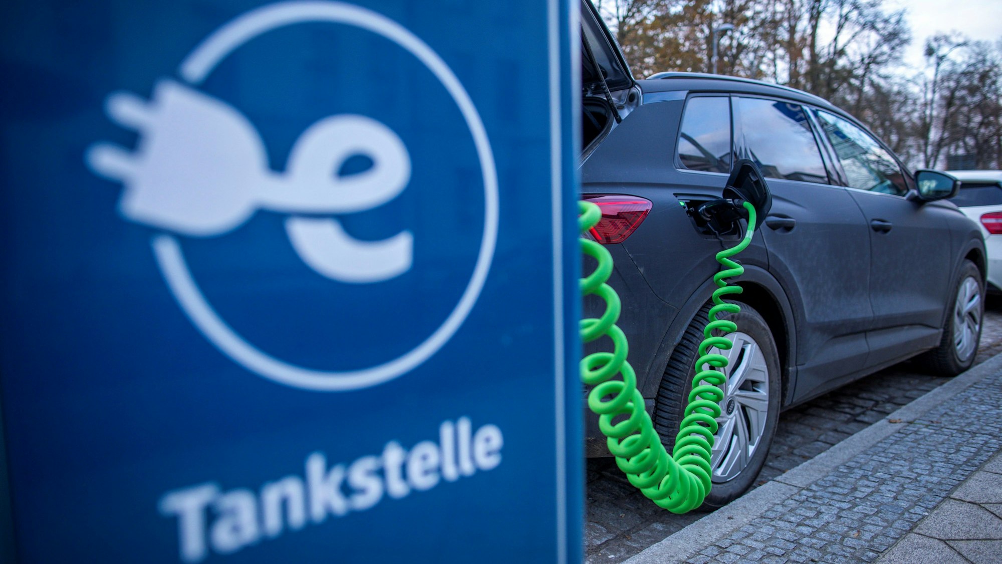 Das Symbolbild zeigt ein Elektrofahrzeug, das an einer blauen Ladesäule mit der Aufschrift „Tankstelle“ steht.