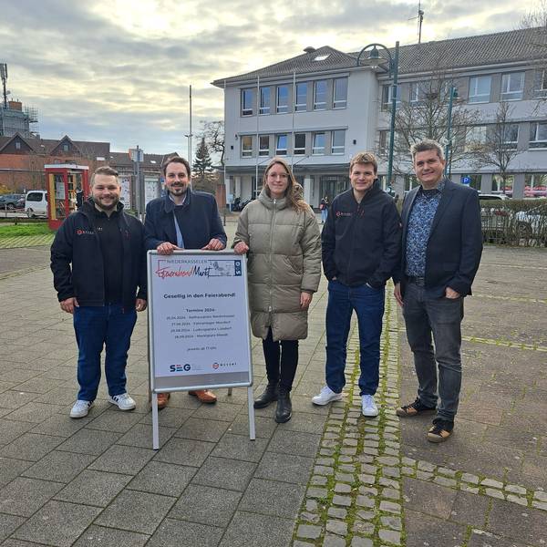 Die Organisatoren der geplanten Niederkasseler Feierabendmärkte stehen auf dem Rathausplatz und präsentieren das Plakat zur Veranstaltungsreihe.