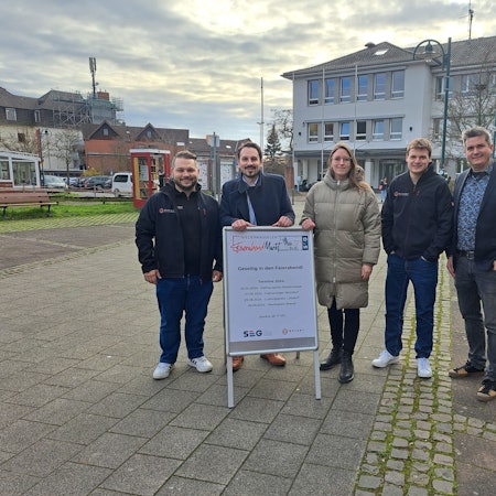 Die Organisatoren der geplanten Niederkasseler Feierabendmärkte stehen auf dem Rathausplatz und präsentieren das Plakat zur Veranstaltungsreihe.