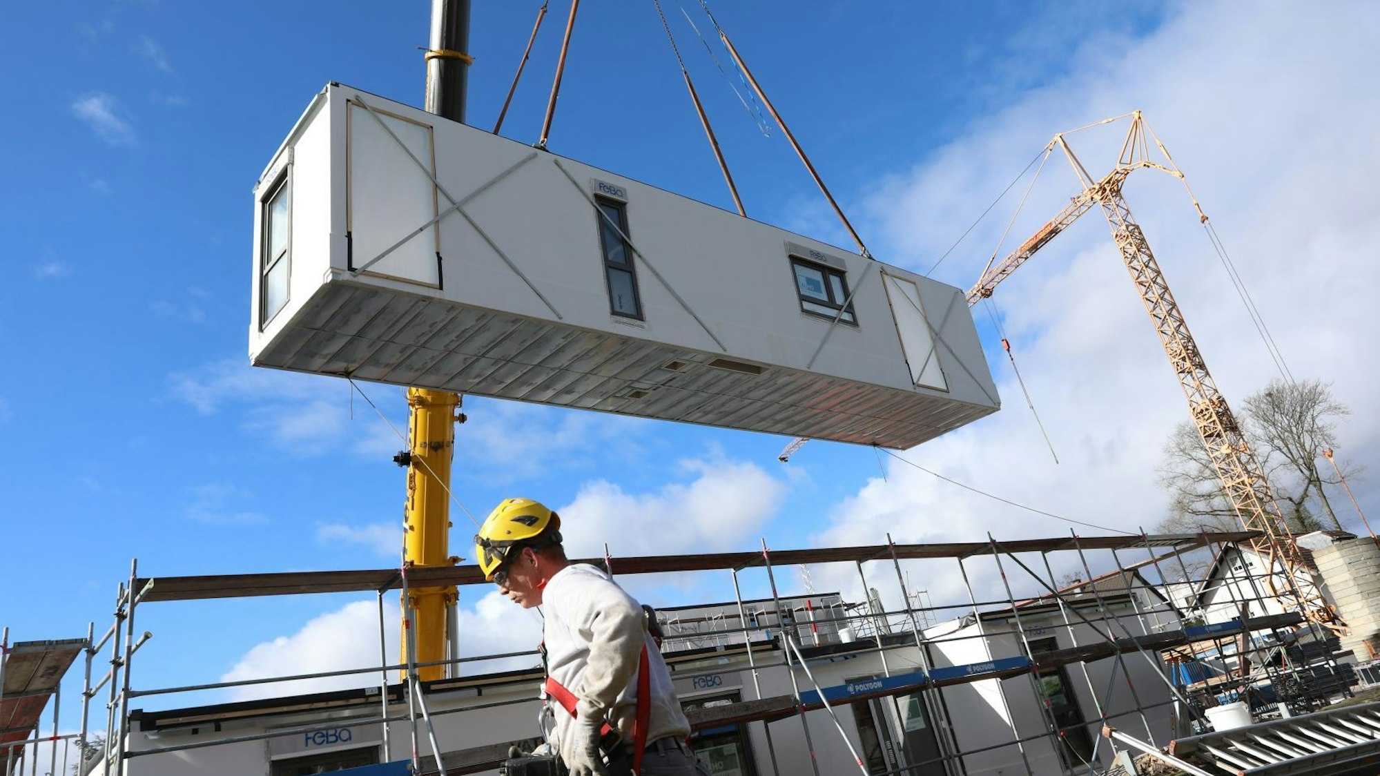 Ein rechteckiges Modul hängt an einem großen Baukran, im Vordergrund steht ein Bauarbeiter.