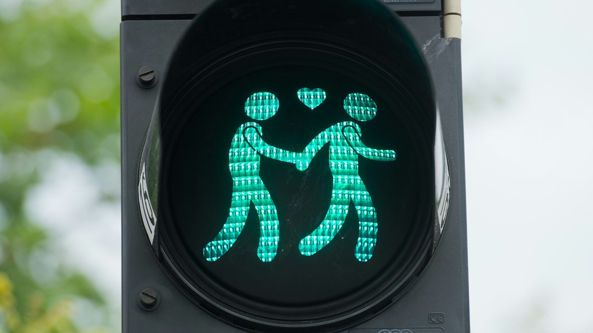 Auf einer Ampel sind zwei Ampelmännchen zu sehen, die Händchen halten. Zwischen ihnen lechtet ein Herz.