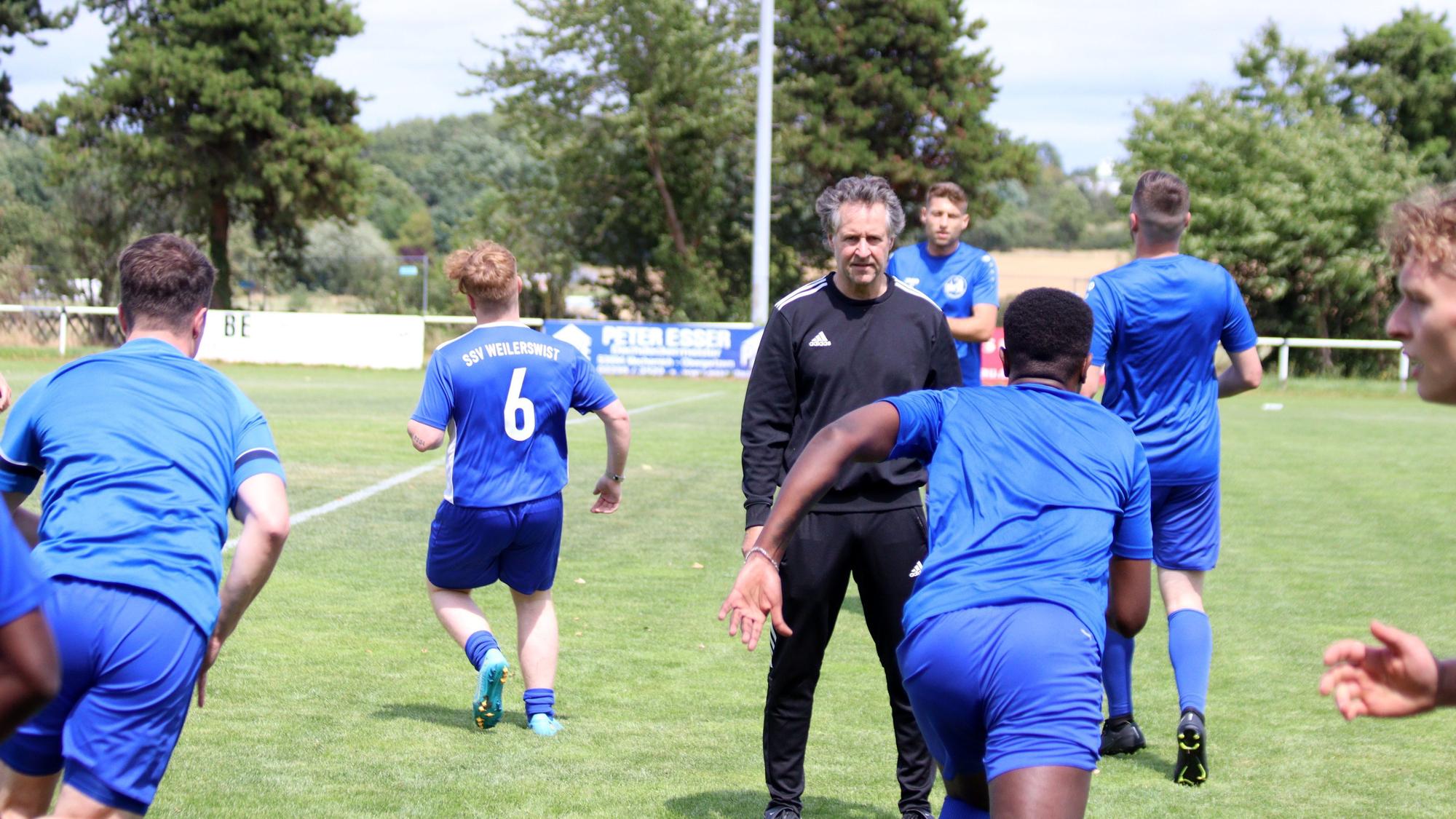 Trainer Horst Bartz steht auf dem Fußballplatz und beobachtet seine Spieler, die Laufübungen absolvieren.